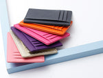 Des porte-cartes en cuir de couleur différente sont regroupés et posés sur un cadre de couleur bleu ciel.