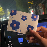 En premier plan, on observe un porte carte avec es fleurs bleues sur un cuir blanc et en deuxième plan, une rue floutée..