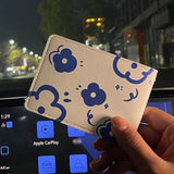 Une main de femme tient un porte carte blanc avec des fleurs bleues et en second plan, on voit le tableau de bord d'une voiture et une rue.