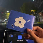 On voit un porte carte en cuir bleue avec une fleur blanche en son centre tandis qu'on peut observer une rue en soirée en deuxième plan.