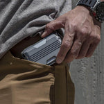 On aperçoit le porte-cartes en métal qui sort de la poche d'un pantalon masculin, tenu par une main qui porte une montre.